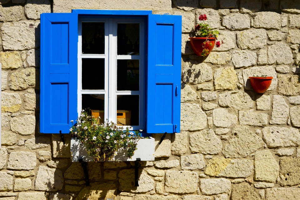 Façade en pierre traditionnelle avec fenêtre à volets bleus ouverts, encadrement blanc, jardinière avec plantes fleuries sur rebord